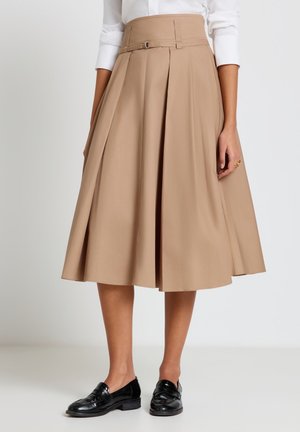 Donna che indossa una gonna midi beige a linea A con vita alta e dettaglio fibbia, abbinata a mocassini neri e una camicia bianca.