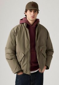 Ung man med en brun keps, olivgrön zip-jacka, vinröd hoodie och mörkblå jeans, stående med händerna i fickorna.