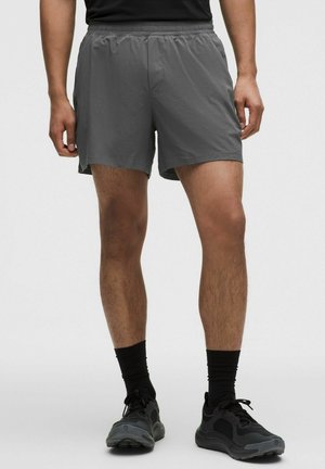 Mand iført grå træningsshorts, sorte mellemlange sokker og sorte løbesko, stående mod en ensfarvet baggrund.