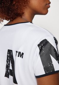 Hvid t-shirt med sort tekstureret tryk, der har store bogstaver på ryggen. Kraven og ærmerne har en kontrastfarvet sort kant.