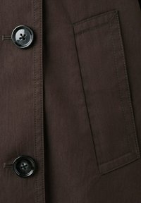 Primer plano de tela marrón oscuro con dos botones negros y un bolsillo rectangular cosido en una prenda.