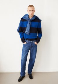 Gestreifter blauer und navyfarbener Pullover, Jeansjacke über den Schultern, blaue Jeans und schwarze Ankle Boots. Angenehme Textur, lässige Passform.