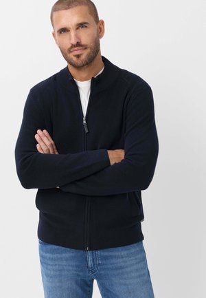 Mann mit kurzen Haaren und Bart, der einen schwarzen Zip-Up-Pullover über einem weißen T-Shirt und blauen Jeans trägt, steht mit verschränkten Armen.