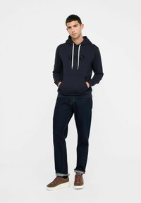 Un jeune homme se tenant avec les mains dans les poches, portant un sweat à capuche bleu marine, un jean foncé et des baskets marron avec des semelles blanches, sur fond blanc.