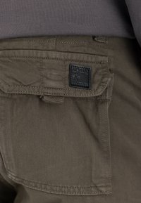 Nærbilde av olivengrønn cargobukselomme med klaff og sort Alpha Industries merke sydd over lommen.