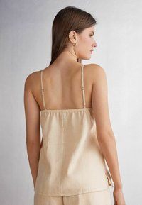 Beige top bez rukávů s tenkými ramínky, elastickým zadním dílem a jemným leskem. Má volný střih a postranní rozparky pro pohodlný pohyb.