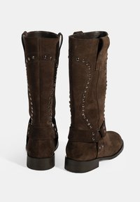 Bottes en suede marron jusqu'au genou avec des studs métalliques décoratifs et un détail de boucle. Présente un bout arrondi et un petit talon.