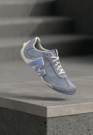 Light grey sneaker with blue accents, suede and synthetic materials, rounded toe, flat laces, textured rubber sole, and prominent side logo.