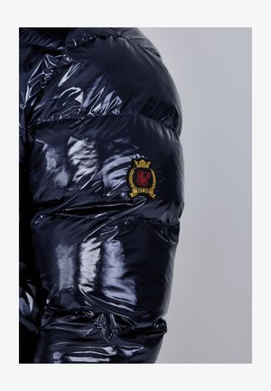 Námořnická puffer bunda s lesklou texturou, zdobená zlatě orámovaným emblémem a příčnými prošívanými sekcemi na rukávu.