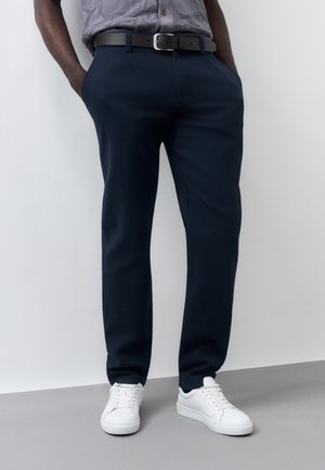 Férfi tengerész kék nadrágban, fekete övvel, szürke inget betűrve és fehér tornacipőben, zsebébe tett kézzel áll egy fehér fal előtt.