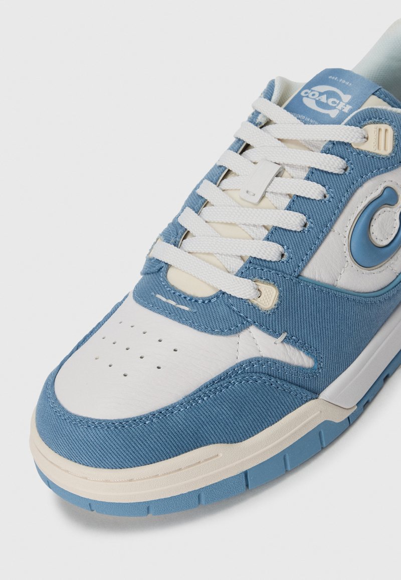 Vit och ljusblå Coach-sneaker med vita snören, texturerat tyg och läderpaneler, samt synlig varumärkeslogotyp på sidan och tungan.