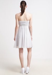 Robe blanche bustier avec un corsage froncé, une jupe évasée et une texture transparente. Portée avec des talons compensés blancs, vue de dos.