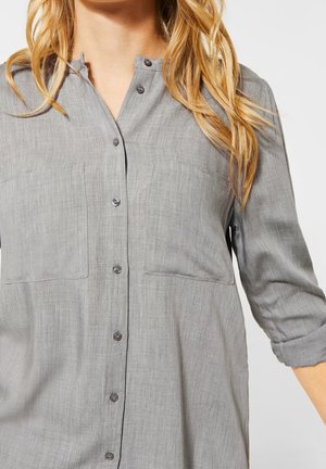 Camicia grigia con colletto a band, due tasche sul petto e maniche arrotolabili. Il tessuto presenta una finitura strutturata con un leggero lucido.