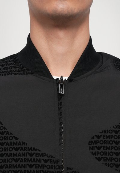 Black bomber jacket with a ribbed collar, featuring a subtle all-over Emporio Armani logo pattern and a central zipper. Smooth texture.