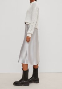 Suéter blanco de punto acanalado con mangas texturizadas, combinado con una falda midi de satén gris claro. Botines negros con paneles elásticos en los costados.