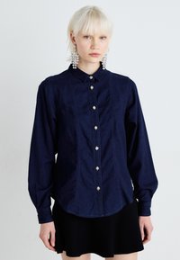 Camicia in denim blu scuro con maniche lunghe, colletto a punta e chiusura frontale con bottoni. Presenta un tessuto testurizzato e dettagli di cucitura sottili.