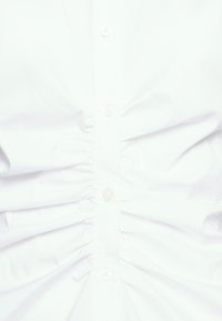 Valkoinen nappipaita, jossa on ryppytys keskellä, valmistettu puuvillakankaasta ja varustettu pienillä, vaaleanvärisillä nappeilla.