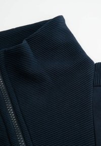 Granatowa bluzka zapinana na zamek z kołnierzem, wykonana z prążkowanej tkaniny. Charakteryzuje się teksturowaną powierzchnią i gładkim zamkiem po jednej stronie, z dopasowanym krojem przy szyi.