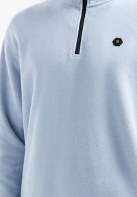 Lichtblauwe pullover trui met een half-rits ontwerp, voorzien van een zwarte rits en een hexagonaal logo-accent op de linker borst. Zachte textuur.
