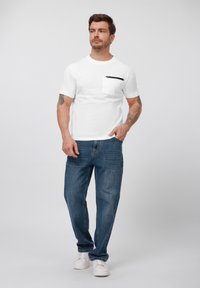 White cotton t-shirt with a chest pocket featuring a black zipper, paired with blue denim jeans. Model wears white sneakers, standing against a light background.