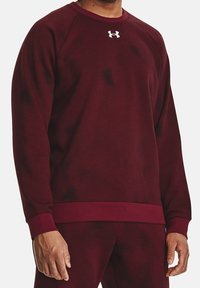 Burgundy atletisk sweatshirt med en texturerad ribbad design, rund halsringning och en kontrasterande vit logotyp på bröstet. Har åtsittande muddar och nederkant.