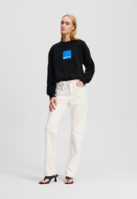 Felpa nera con patch logo blu, abbinata a jeans bianchi a vita alta e sandali neri con punta aperta. Texture lisce, vestibilità comoda.
