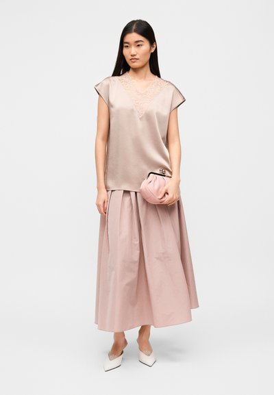 Απαλό ροζ σατέν τοπ με δαντέλα στη λεπτομέρεια, συνδυασμένο με αέρινη, ταιριαστή ροζ φούστα. Το μοντέλο κρατάει ένα απαλό ροζ τσαντάκι και φοράει λευκές μυτερές μπαλαρίνες.