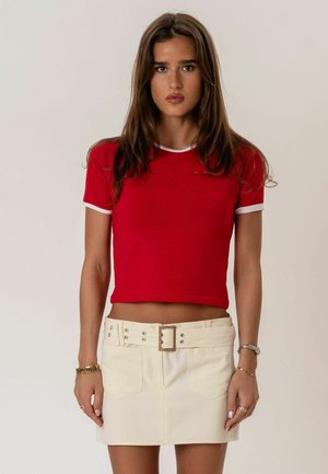 Mlada žena s dugom smeđom kosom u uskoj crvenoj kratkoj majici s bijelim obrubom i bež mini suknji širokog remena.