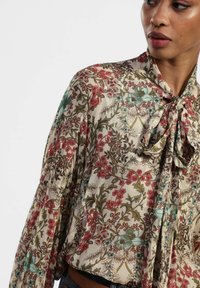 Blusa floreale con sfondo crema, intricati motivi di fiori rossi e turchesi, maniche lunghe e dettaglio del colletto annodato.