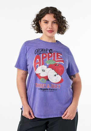 Kvinde med krøllet hår iført en lilla T-shirt med røde æbler og teksten "Økologisk æble sødt og saftigt" på en hvid baggrund.