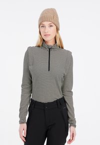 Schwarz-weißer, gemusterter Langarm-Reißverschlussoberteil mit hohem Kragen und slim fit. Kombiniert mit enganliegenden schwarzen Hosen. Beiger Strickmütze.