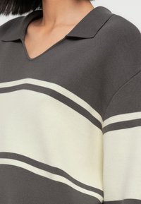 Κοντινό πλάνο ατόμου που φοράει γκρι πουλόβερ με φαρδιές λευκές και στενές σκούρες γκρι οριζόντιες ρίγες και μικρό γιακά.