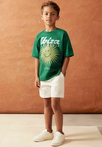 Camiseta verde con gráfico del sol y texto "Ibiza", combinada con pantalones cortos blancos y zapatillas blancas, en un modelo de pie contra un fondo naranja.