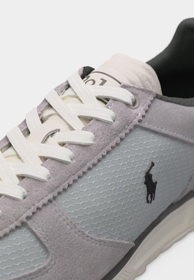 Posnetek iz blizu sive in bele teniske z belimi vezalkami, mrežasto teksturo, semišimi paneli in črnim logotipom igralca polo ob strani.