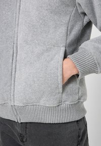 Grijze fleece sweater met geribbelde manchetten en tailleband, voorzien van een zijzak. Het materiaal heeft een zachte, gestructureerde afwerking.