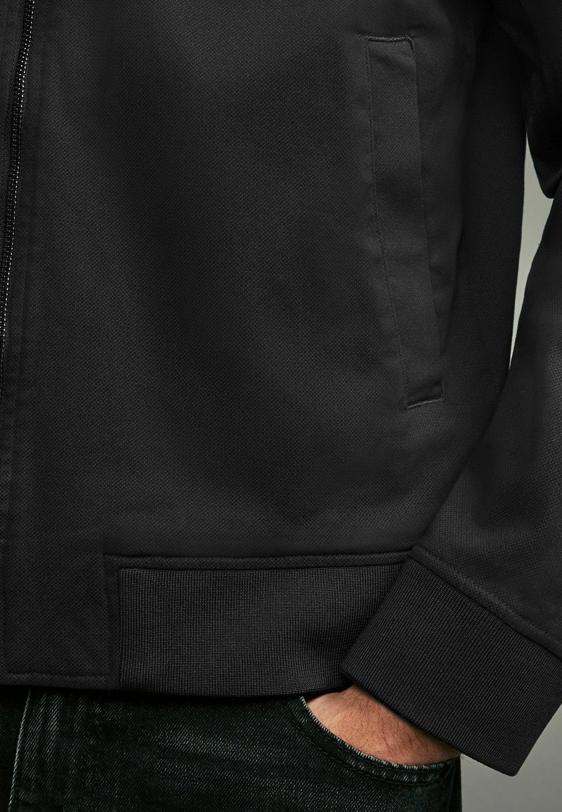 Schwarze Jacke mit strukturiertem Finish, Stehkragen und gerippten Bündchen. Seitentasche auf der linken Seite, über dunkler Denim getragen.