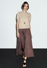 Una modella si trova davanti a uno sfondo neutro, indossando un top beige e una gonna marrone fluida, accessoriata con una lunga collana e sandali neri.