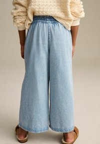 Világoskék, bő patkós farmernadrág, gumis derékkal, farmer anyagú textúrával és foszlott szegéllyel, barna slip-on cipőkkel párosítva.