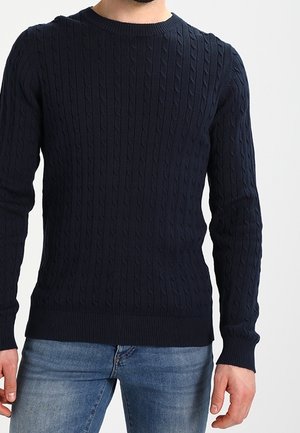 Man wearing a fitted navy blue cable knit sweater and blue jeans, shown from the neck to hips against a plain white background.