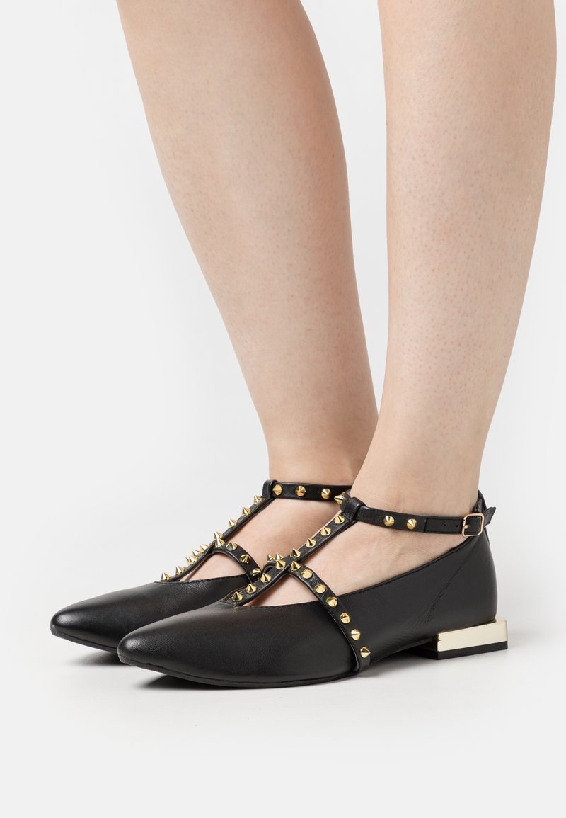 Chaussures plates noires à bout pointu avec brides à la cheville et clous dorés, portées sur des jambes à peau claire sur fond blanc.