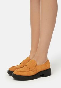 Mocasines de ante en un vibrante color naranja con una suela negra gruesa. Presentan una amplia correa en la parte delantera y un diseño de punta redondeada.