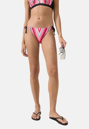 Frau trägt mehrfarbigen gestreiften Bikinihöschen, hält Sonnenbrille in der rechten Hand und trägt schwarze Flip-Flops an nackten Füßen.