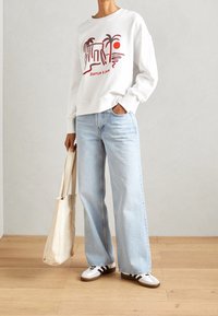 White sweatshirt with a graphic design featuring palm trees and a sun, paired with light blue wide-leg jeans and white sneakers.