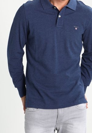 Homme portant un polo bleu marine à manches longues avec le logo GANT sur la poitrine et un pantalon gris clair, mains dans le dos.