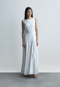 Lang, hvid, ærmeløs kjole med rundet halsudskæring, tætsiddende overdel og udvidet, lagdelt nederdel. Glat stoftekstur, minimalistisk design.