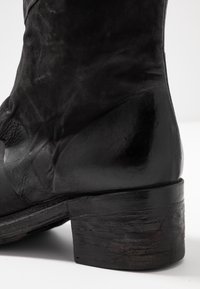Bota de cuero negro con un acabado suave, punta puntiaguda y un tacón robusto y texturizado. Presenta un diseño elegante y ajustado.