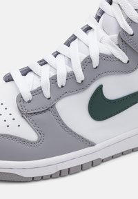 Nærbilde av en grå og hvit sneakersko med hvite lisser og en mørkegrønn Nike swoosh-logo på siden.