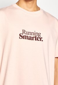 Camiseta de manga corta, de color rosa claro, confeccionada en tela suave y con el texto "Corriendo de manera más inteligente" en un tipo de letra serif marrón oscuro en la parte delantera.