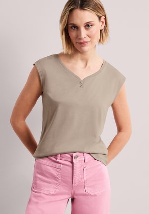 Kvinde med kort blondt hår iført en ærmeløs beige top med to knapper og højtaljede lyserøde bukser, stående med hænderne bag ryggen.