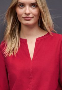 Mujer rubia con piel clara, vistiendo un top rojo de cuello en V, mirando hacia adelante con una expresión neutral frente a un fondo gris oscuro.
